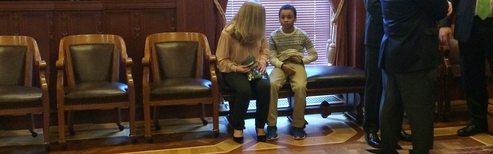 Female speaking with a boy sitting on formal chairs in a legislative building with men in suits standing to the right.