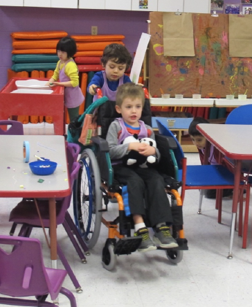 Child pushing another child in a wheelchair holding a stuffed animal