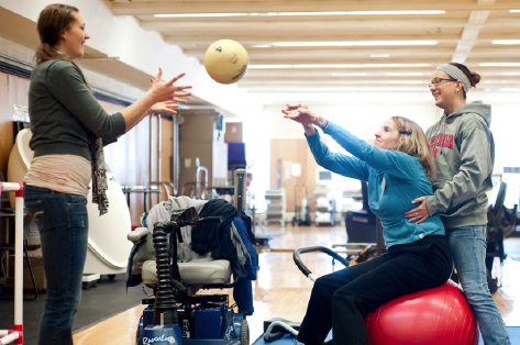 Adult woman reaching her arms out to catch a ball thrown by a woman sitting on an exercise ball supported by another woman in an indoor gym.