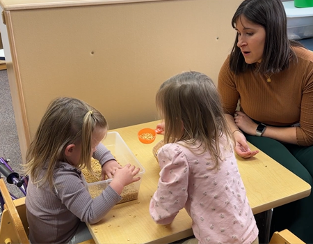 Two young girls playing with a bin of dry pasta with a women sitting at the table talking with them